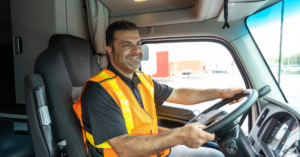 Truck driver wearing a high-visibility vest smiling while driving, showcasing the use of ISAAC Coach technology.