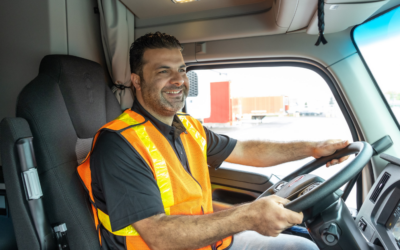 Truck driver wearing a high-visibility vest smiling while driving, showcasing the use of ISAAC Coach technology.