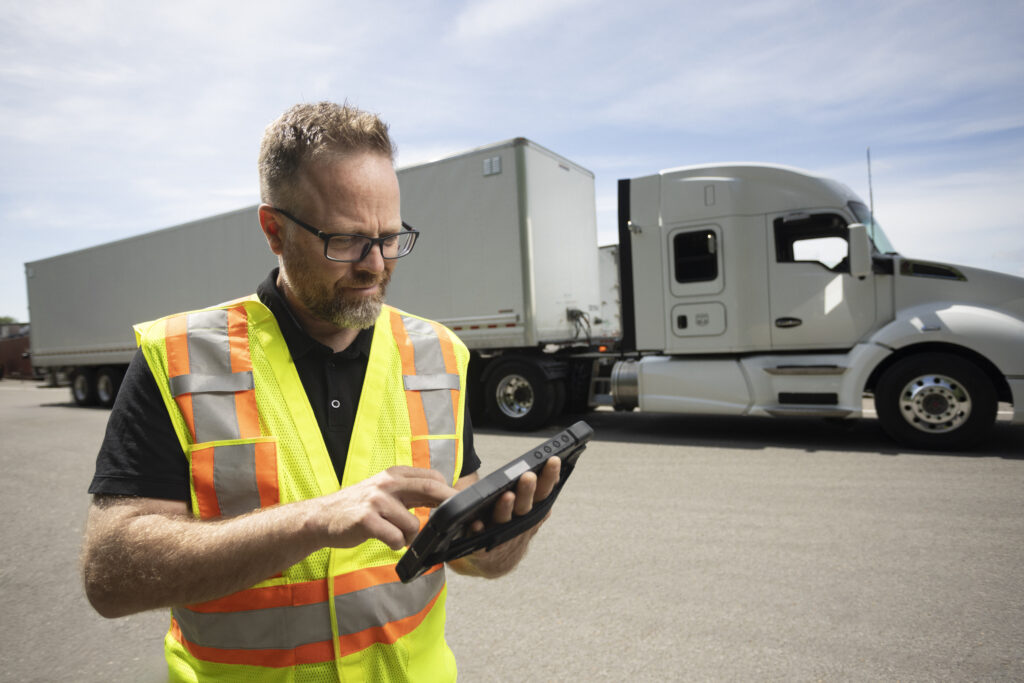 Chauffeur de camion portant un gilet de sécurité utilisant une tablette dans un parking avec un semi-remorque blanc derrière lui.