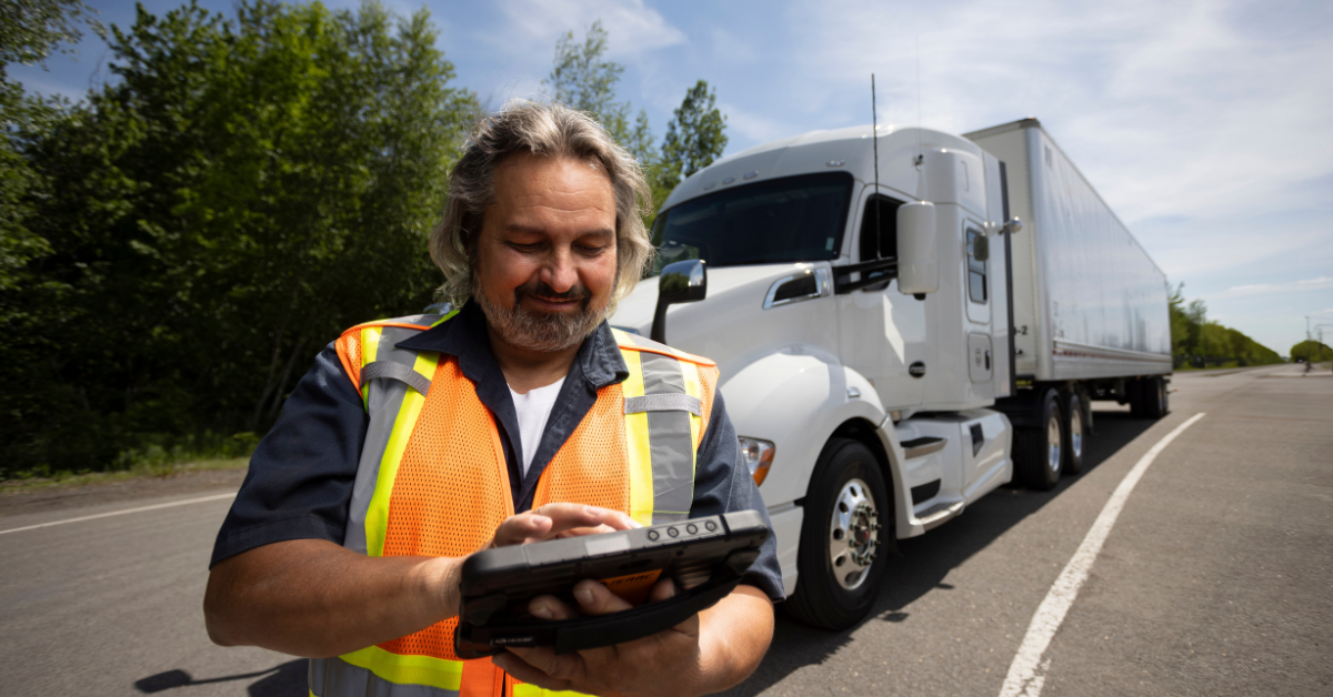 Truck driver wearing a safety vest using a tablet roadside with a white semi-truck in the background.