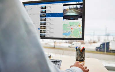 Fleet management software dashboard showing real-time vehicle tracking data with location map and video feed, displayed on a monitor in an office with a blurred highway view in the background.