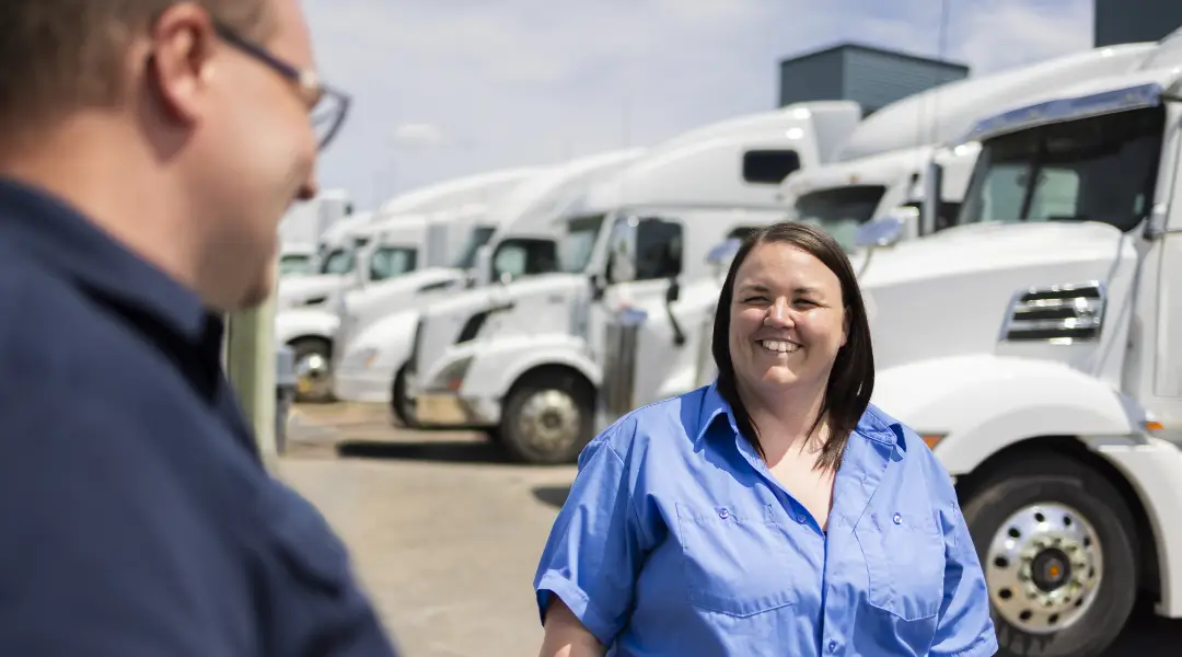 A smiling female fleet manager wearing a blue shirt, speaking with a colleague, with a row of semi-trucks in the background.