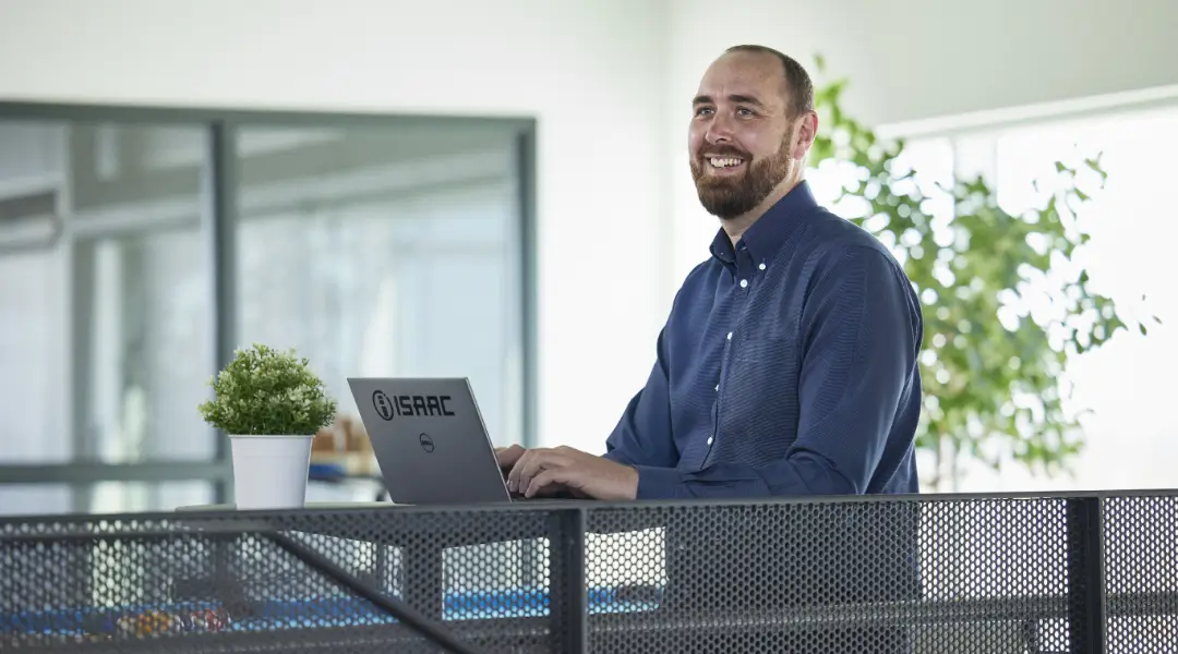 A bearded professional in a blue shirt smiling while working on an ISAAC-branded laptop in a bright, modern office.