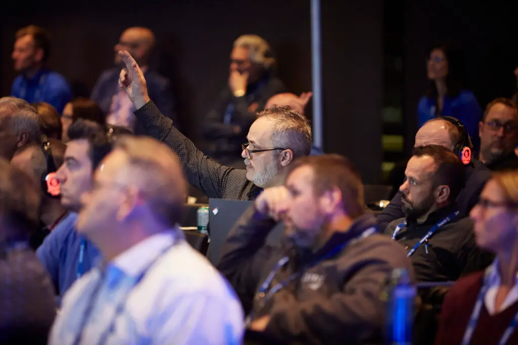 Attendee raising hand to ask a question during a logistics user conference, surrounded by engaged audience members